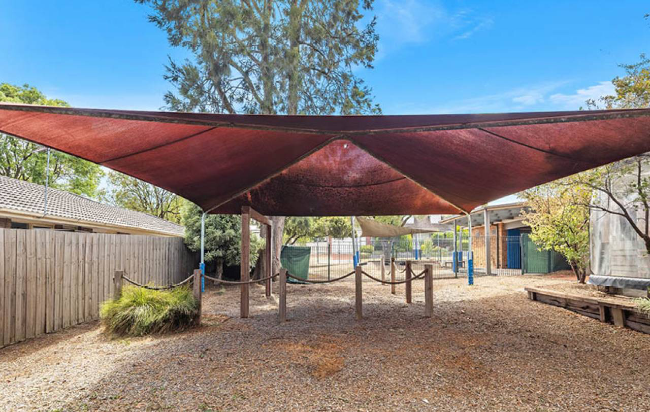 Red shade sail on timber posts over a mulched play area, with fencing and neighbouring rooftops beyond.